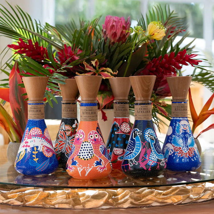 Set of colorful bottles with artistic designs on a table with a floral arrangement in the background.